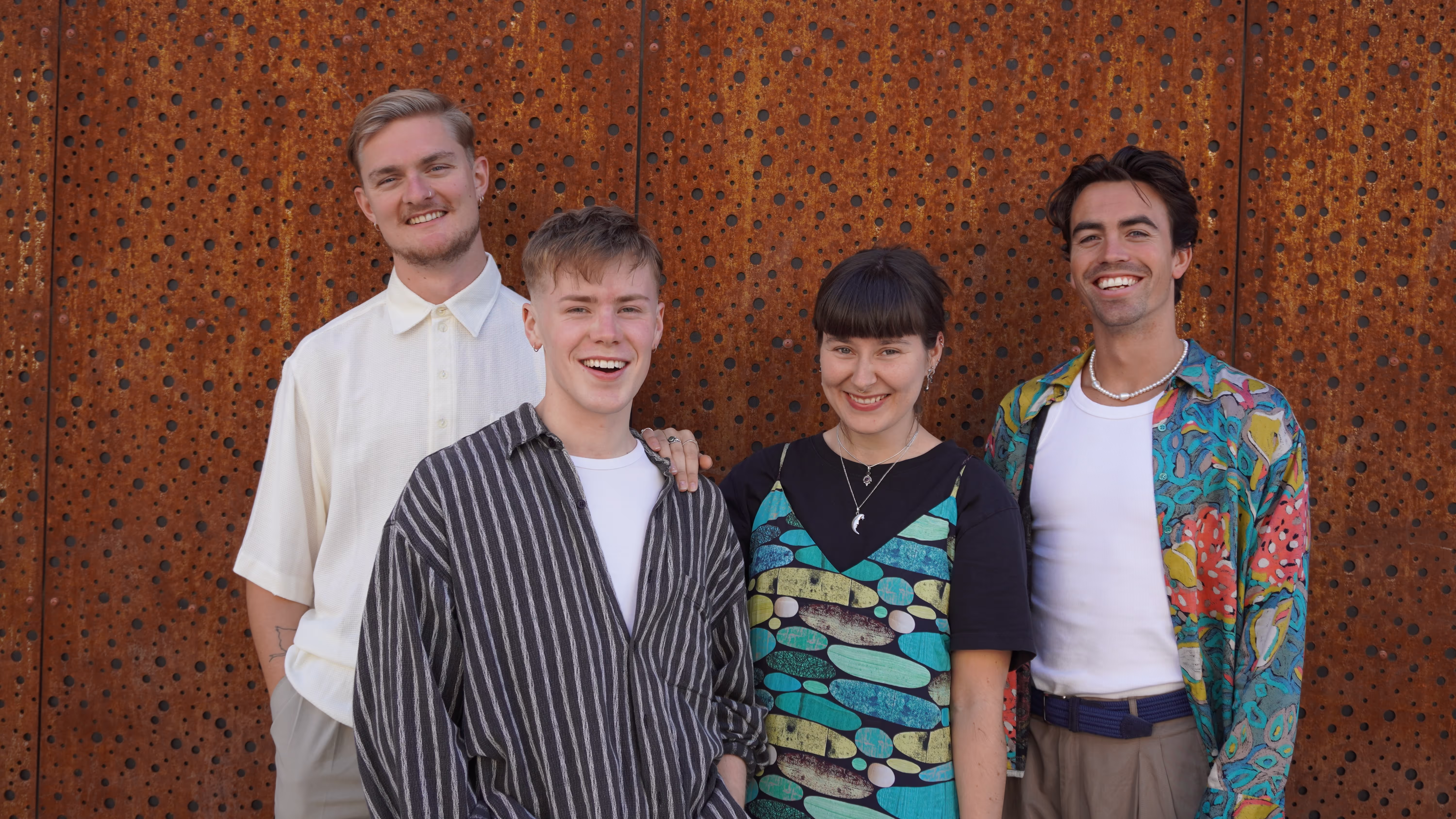 Fire unge mennesker står foran en rustrød bakgrunn. De har på fargerike klær og smiler fint til kamera.