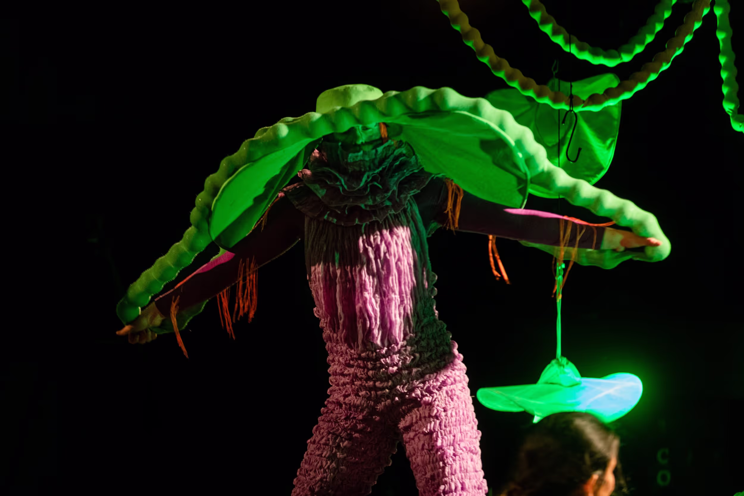 Et bilde fra forestillingen Himstegrimser og andre duppeditter. Et menneske i rosa drakt står med noe grønt på hodet sitt.