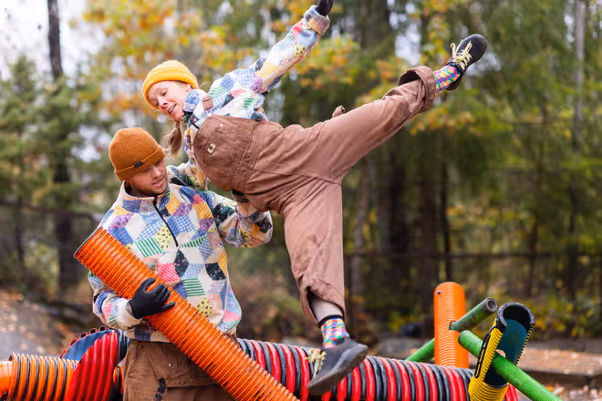 Mange fargerike rør er satt sammen til en konstruksjon. 2 dansere strekker seg foran en skog