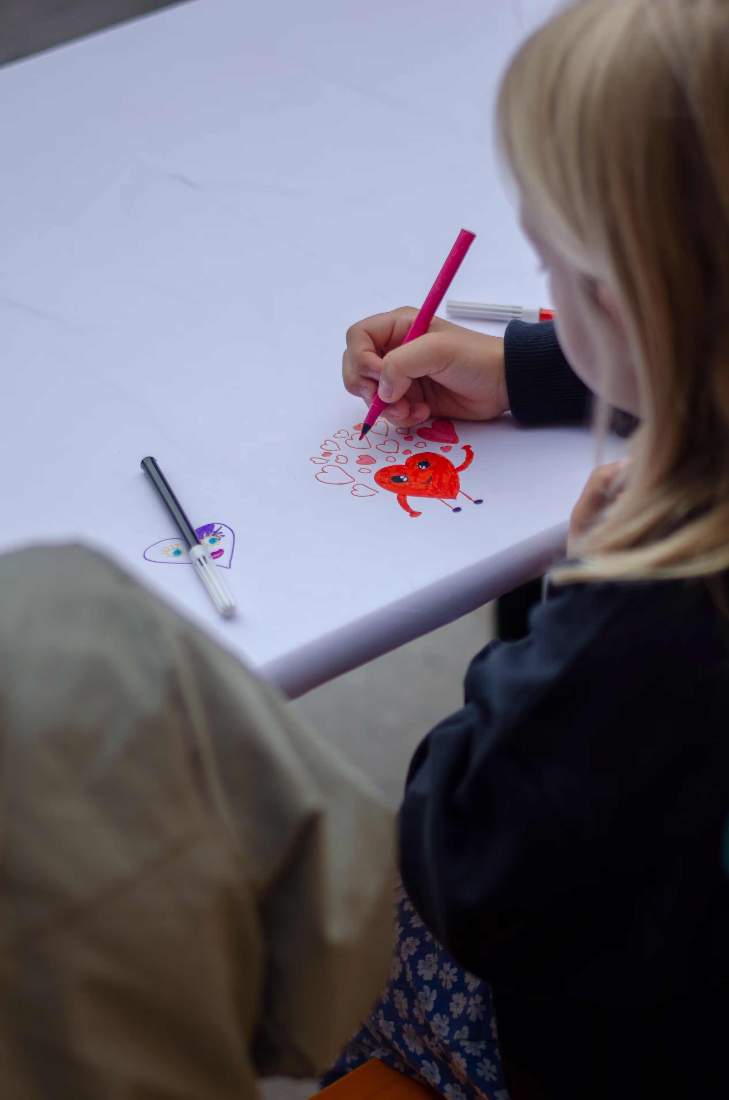 Over skulderen til to barn ser vi at de tegner et hjerte sammen på et ark.